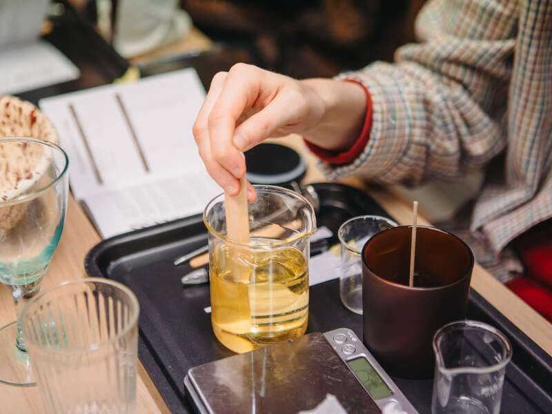 A close up of a person stirring melted wax in a jar, with candle making equipment around